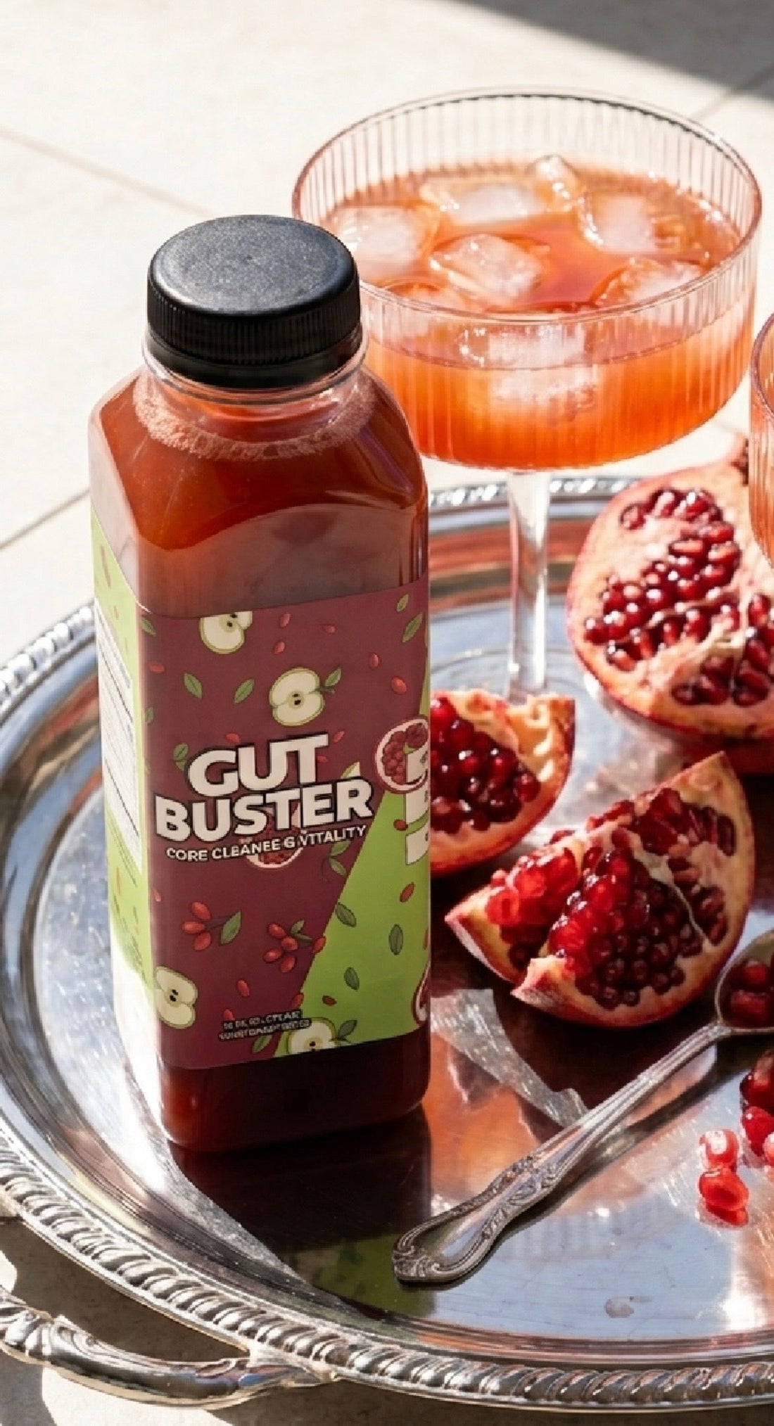 Gut Buster gut-support drink bottle on a silver tray with a pomegranate cocktail and fresh pomegranate